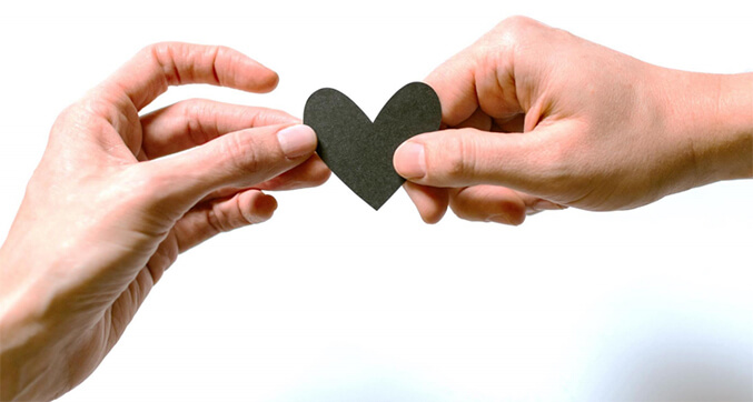 A black heart-shaped cutout is cupped in the hands of a man and a woman. Their hands meet in the center of the heart, suggesting a bond or connection.
The heart is made of paper and appears flat and two-dimensional.
The couple’s hands are lightly tanned, with manicured fingernails. The woman’s nails are painted a light pink color.
The background is blurry and appears light-colored.